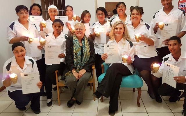 Die tuisversorgers met hul brandende lampies â€“ voor van links: Celeste Dunsdon, Sanna Speelman (beste prestasie: strykwerk), Suster Irene de Waal, El-Marie Fourie (hoogste lof vir teorie en prakties) en Franklin Eksteen (eervolle vermelding).  Agter: CornÃ© Dunsdon (beste blommerangskikking), Trudie Cupedo, Elna Kondokter (prakties en huishouding met lof), Rhowina Kammies, Magdalena Arends (naaldwerktoekenning), Nicole Adonis, Jocoline Louw (slaag prakties met lof), Marlene Gertse (klaskaptein â€“ slaag prakties en tuisversorgingsteorie met lof) en Peter-Lee Karelse (slaag prakties met lof).